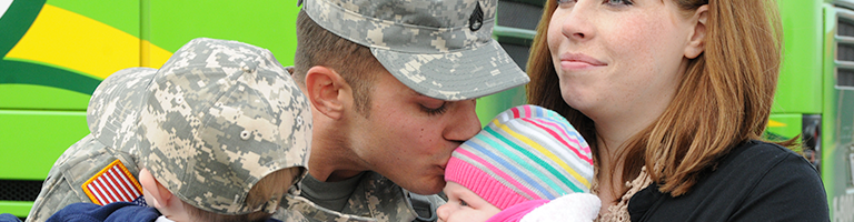 Military Family greets father. Dad kisses infant daughter while mom smiles.
