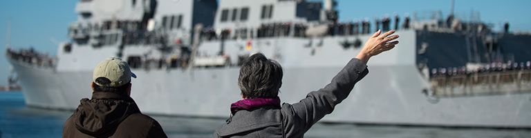 Military parent wave to Navy ship from the coastline.
